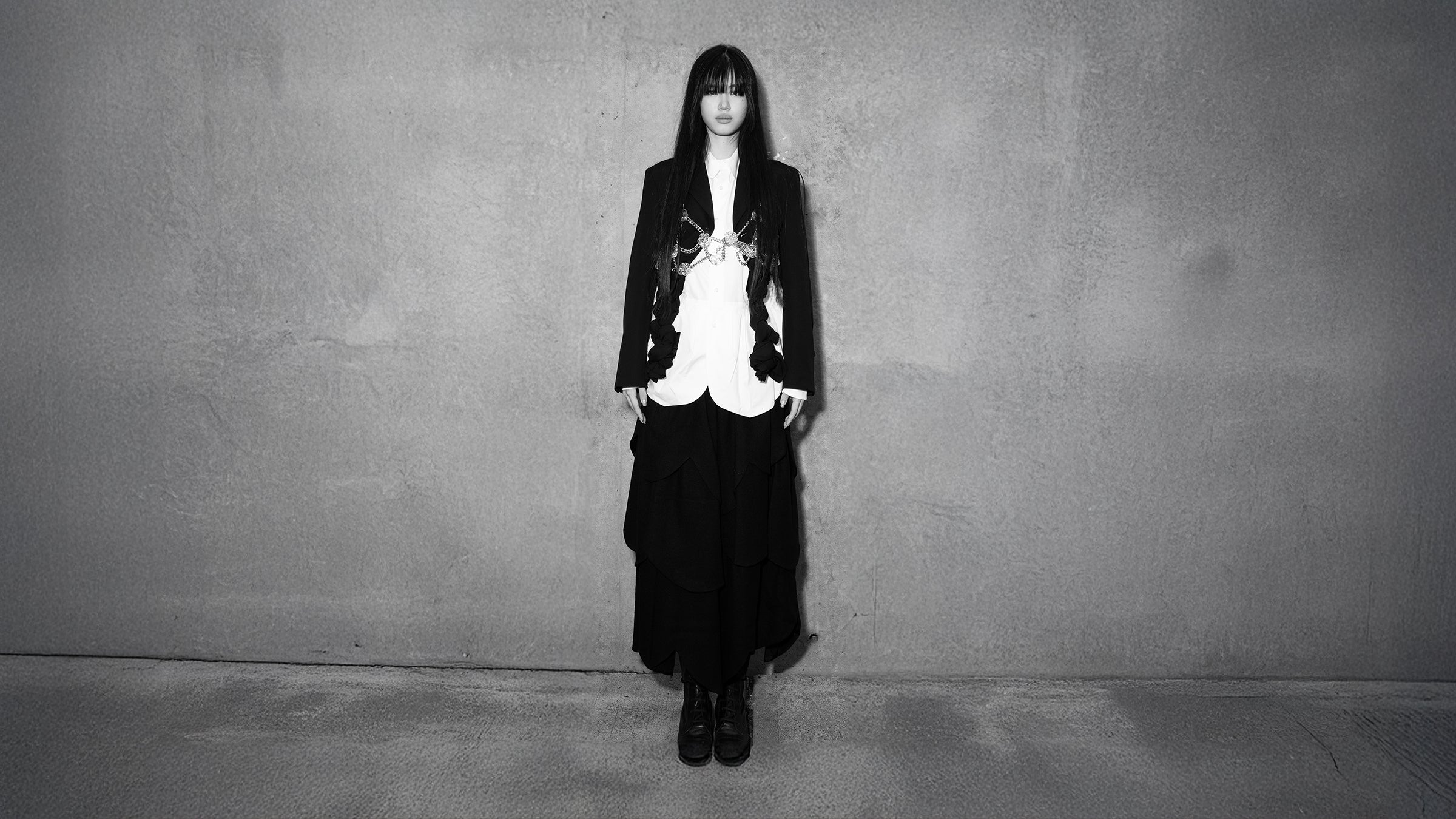 Model wearing a black layered skirt and tailored blazer with a white button-up shirt and chain detail from Comme des Garçons, photographed against a concrete wall for Atelier New York’s Fall/Winter 2025 sale
