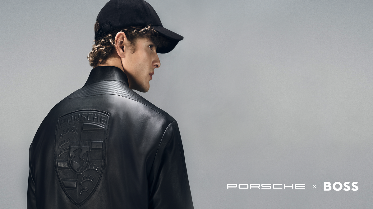 A man shown from the back wearing a black leather jacket with an embossed Porsche crest on the back, a black baseball cap, and a Porsche x BOSS logo is visible in the bottom right corner against a gray studio background.
