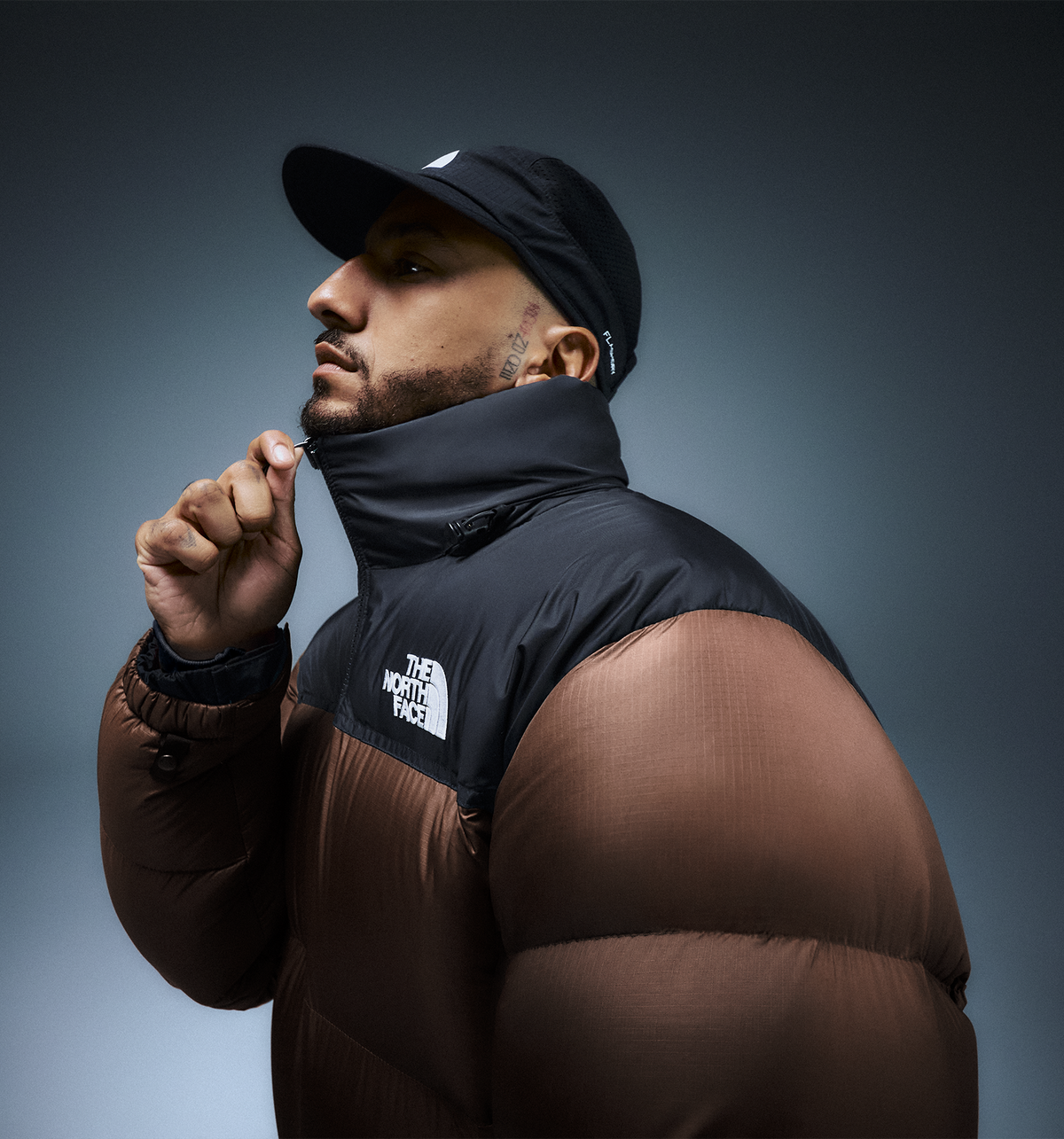 A man wearing a brown and black puffer jacket with a white The North Face logo on the chest, paired with a black cap, posed against a dark gray studio background.