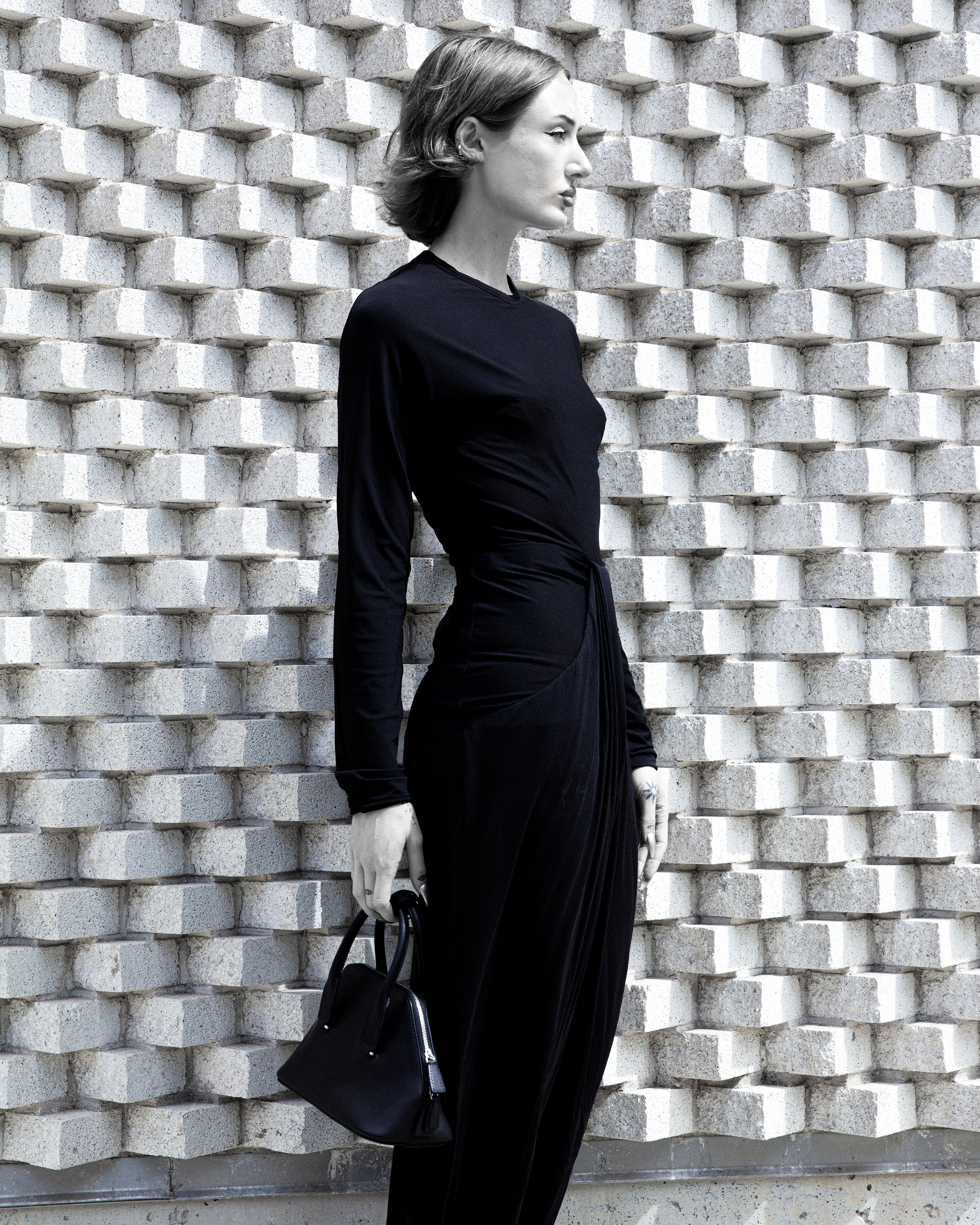 Female model wearing a black long-sleeve Anselm Dress and carrying a structured blackhandbag by The Row, photographed against a geometric stone wall for Atelier New York’s Women’s Collection.