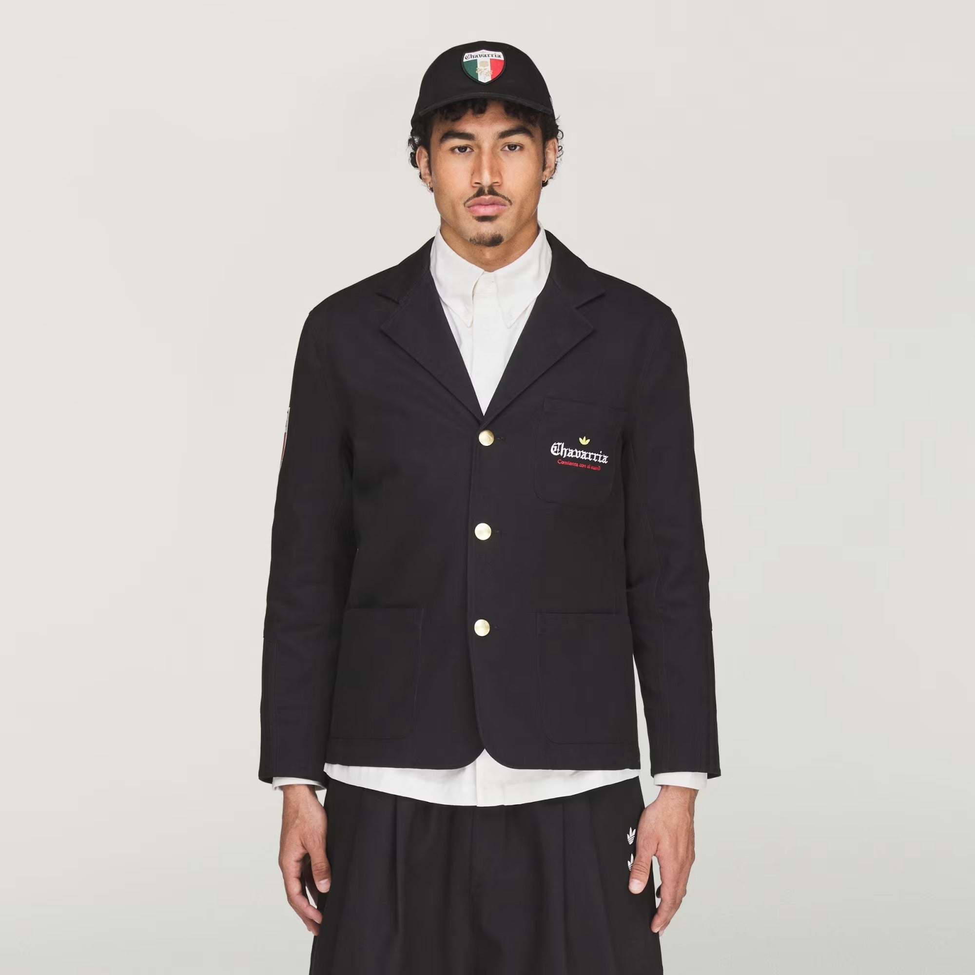 Person wearing a black blazer with a logo, white shirt, and black cap on a light gray background