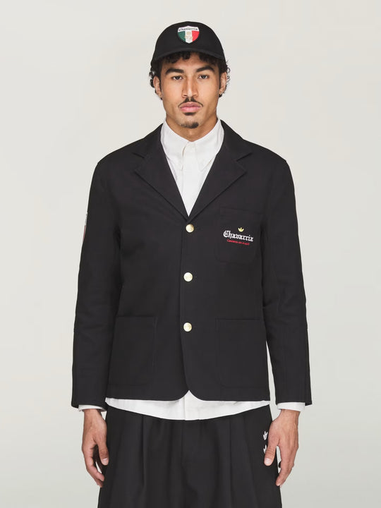 Person wearing a black blazer with a logo, white shirt, and black cap on a light gray background